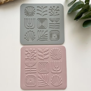 Può includere: Due sottobicchieri quadrati con un design botanico. Uno è grigio, l'altro rosa. Ogni sottobicchiere presenta nove forme in rilievo, tra cui fiori stilizzati, foglie e motivi geometrici. I sottobicchieri sono realizzati in materiale flessibile.
