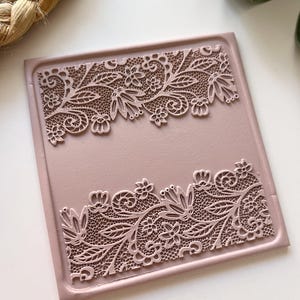 May include: A square, dusky pink decorative tray featuring detailed lace-style floral and leaf patterns embossed along the top and bottom edges. The tray has a raised border and a smooth surface, ideal for displaying small items.