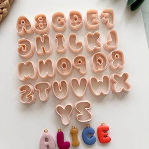 May include: A set of light peach-coloured plastic alphabet letter cookie cutters arranged in rows, with five individual colourful letter charms spelling the name ALICE positioned below, each featuring a small gold-toned metal jump ring at the top.