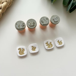 Peut inclure: Ensemble de tampons et d'ornements carrés sur le thème de l'écureuil. Les tampons présentent des motifs d'écureuils gris sur des bases rondes en bois avec des manches roses. Les ornements carrés présentent des motifs d'écureuils dorés sur fond blanc.