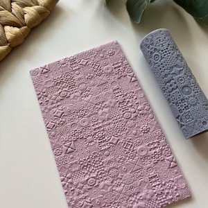 Puede incluir: Un juego de dos herramientas de relieve de silicona púrpura con un patrón geométrico. La herramienta más grande es una lámina plana y la herramienta más pequeña es un rodillo.