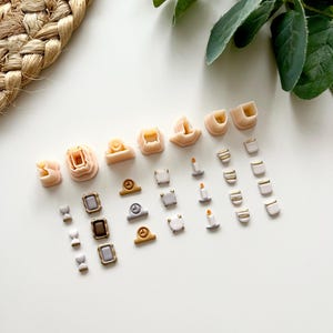 May include: A selection of small polymer clay charms in various shapes and colours. The charms include miniature shoes, clocks, candles, and picture frames. The colour palette is primarily neutral, with accents of gold and silver.