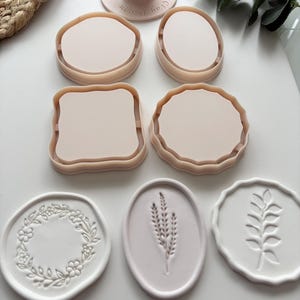 Puede incluir: Un conjunto de cortadores de arcilla rosa claro y formas de arcilla blanca en relieve a juego. Los cortadores incluyen formas circulares, ovaladas y cuadradas festoneadas. Un cortador presenta el texto "Vie Clay Design" en su mango. Las formas en relieve muestran patrones de corona floral, espiga de trigo y hojas.