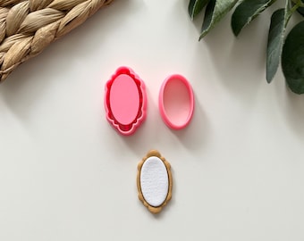 Puede incluir: Dos cortadores de galletas ovalados de plástico rosa y una galleta ovalada dorada con un centro blanco.