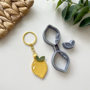 Puede incluir: Un llavero de cuero amarillo en forma de limón con hojas verdes y un anillo de llave dorado. El llavero está al lado de un cortador de galletas de limón de plástico azul.