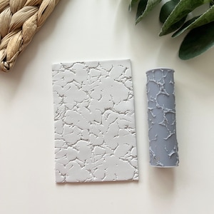 May include: A white rectangular textured tile and a grey cylindrical textured roller. The tile and roller have a cracked, weathered appearance. These items are likely used for creating textured surfaces in crafts or art projects.