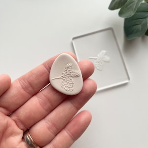 May include: A white ceramic oval shaped stamp with a detailed design of a pine cone and branch.