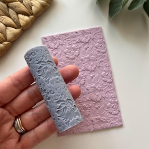 May include: A gray rolling pin with a floral design, next to a pink sheet of fondant with the same floral design embossed on it.
