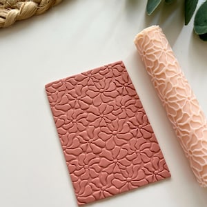 Puede incluir: Un sello rectangular de arcilla y un rodillo a juego, ambos de color terracota, con un diseño floral. El sello es plano, mientras que el rodillo es cilíndrico. Los artículos se muestran sobre una superficie blanca.