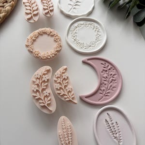 May include: A collection of pale pink and white clay stamps and corresponding embossed impressions featuring botanical designs including floral wreaths, crescent moons, and sprigs of leaves. The set includes various shapes such as circles, ovals, and crescents with intricate, raised floral patterns for crafting.