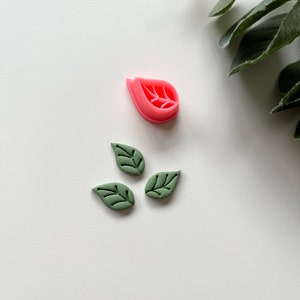 May include: A set of three green leaf-shaped fondant cutters and a pink flower-shaped cutter. The cutters are on a white surface.