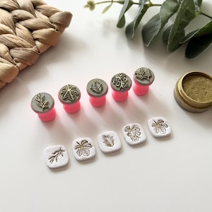 May include: A collection of decorative stamps with botanical designs, including leaves and floral patterns. The stamps have gold-colored details and are mounted on pink bases. A small container of gold ink is also present.