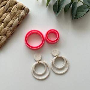 May include: A pair of white double-circle earrings with a textured finish. The earrings have a gold hook closure. The earrings are displayed on a white surface with pink plastic circles.