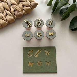 Puede incluir: Un conjunto de seis sellos grises y dorados con diseños botánicos y de mariposas. Los sellos están dispuestos con un cuadrado de cuero verde que tiene diseños en relieve a juego. Una cesta tejida y vegetación están en el fondo.