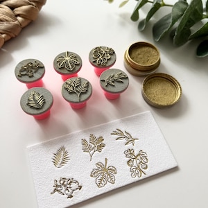 May include: A collection of leaf-themed stamps with pink handles and gray tops. The stamps are arranged with a small container of gold ink and a white card with stamped leaf designs. The stamps and ink are for crafting.