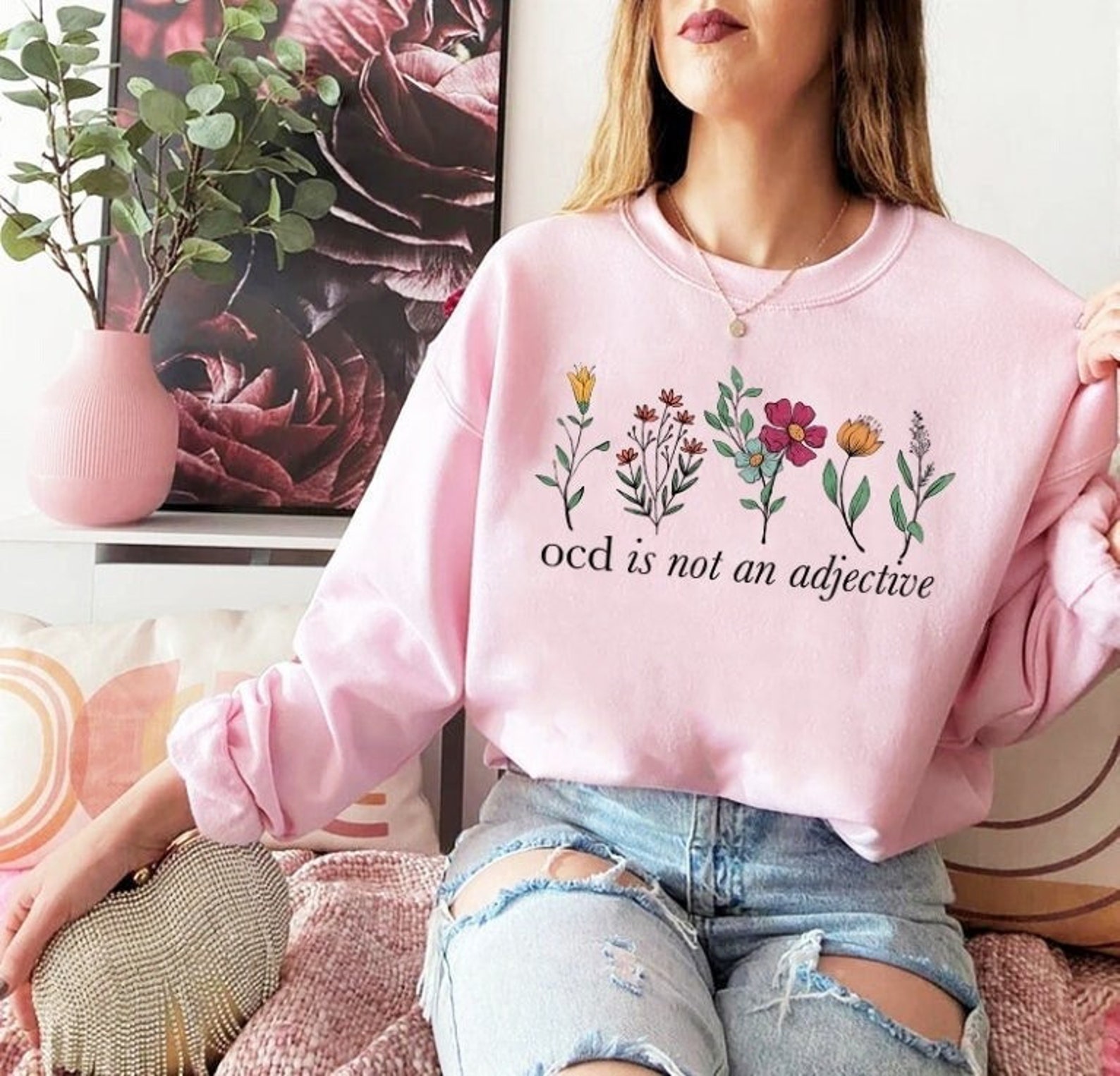 Woman sits on sofa in feminine surroundings while wearing pink sweatshirt with flowers that says "OCD is not an adjective."