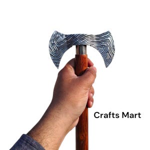 Puede incluir: Un hacha de doble filo con un mango de madera. La cabeza del hacha es de metal plateado con un diseño estampado. El mango es de madera marrón. El hacha está sostenida en la mano de una persona. Crafts Mart está escrito en texto negro sobre un fondo blanco.