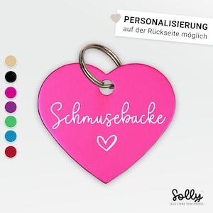 Könnte beinhalten: Rosa herzförmiges Metallschild mit weißer Aufschrift "Schmusebacke" und einem kleinen weißen Herz. Das Schild hat einen silbernen Ring oben.