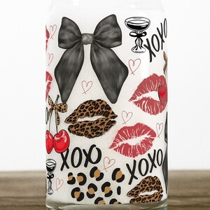 Op de afbeelding: Een doorzichtige glazen beker in de vorm van een blikje met een witte binnenkant, versierd met een zwarte strik, rode lippen, lippen met luipaardprint, kersen, champagneglazen, hartjes en de tekst "xoxo".