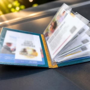 Puede incluir: Un tarjetero de cuero color verde azulado con lomo amarillo y múltiples fundas de plástico transparente. El interior contiene varias tarjetas y documentos borrosos insertados en los bolsillos, mostrando un diseño estructurado para organizar tarjetas de identificación.