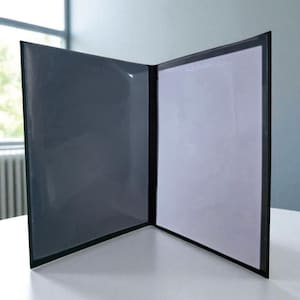 Op de afbeelding: Een open, zwarte presentatiemap met een transparante plastic overlay aan de linkerkant en een witte papieren inleg aan de rechterkant. De map staat op een wit oppervlak, met een raam en een radiator op de achtergrond.