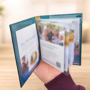 Puede incluir: Un tarjetero de color azul cerceta con aspecto de cuero y acabado brillante, que cuenta con varias fundas de plástico transparente para organizar tarjetas. El interior contiene varias tarjetas blancas con texto impreso e imágenes borrosas.
