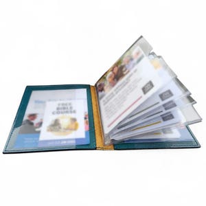 Puede incluir: Un tarjetero de cuero color verde azulado con varias fundas de plástico transparente que contienen tarjetas impresas. La tarjeta frontal presenta el texto "FREE BIBLE COURSE" y "GO TO JW.ORG". El lomo interior es de un color beige contrastante.