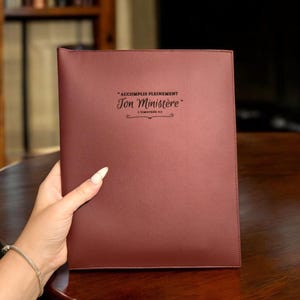 May include: A burgundy leather portfolio with the text "Accomplis pleinement Ton Ministère" in black script. The portfolio is held by a person. The text is from 1 Timothy 4:5.