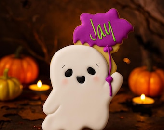 May include: A ghost-shaped sugar cookie decorated with white icing, featuring a smiling face with black eyes and pink cheeks. The ghost holds a purple balloon-shaped cookie with the name Jay written in bright green icing.