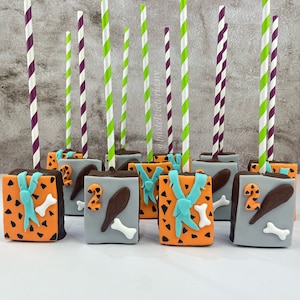Puede incluir: Un conjunto de 10 cake pops decorados con un tema de dinosaurio. Los cake pops están cubiertos de fondant en naranja, marrón, gris y azul. Los cake pops están en palos y tienen pajitas rayadas de morado y verde.