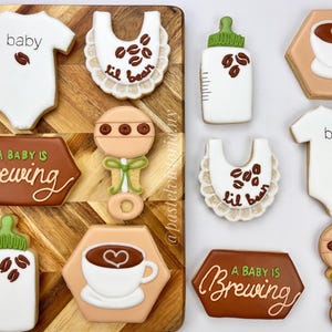 Op de afbeelding: Assortiment van baby-thema koekjes op een houten plank. Koekjes zijn onder meer een witte romper met "baby" en een koffieboon, slabbetjes met "lil bean", babyflessen, koffiekopjes, een rammelaar en een bord met "A Baby Is Brewing".