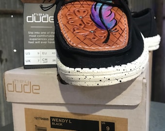 Puede incluir: Un zapato sin cordones negro Hey Dude Wendy L con un panel de cuero sintético tostado que presenta flores en relieve y una pluma pintada en azul y morado. El zapato tiene una suela blanca con motas y una etiqueta negra con el texto Hey Dude.