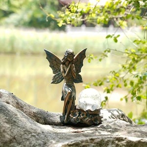 May include: A bronze-colored fairy statue with detailed wings and a serene expression. The fairy stands beside a cracked glass orb, set on a base with floral accents. The statue is placed on a weathered wooden surface, with a blurred natural background.