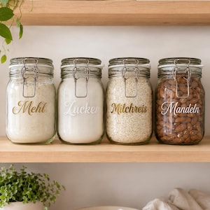 Könnte beinhalten: Vier Gläser mit Metallverschlüssen stehen auf einem Holzregal. Die Gläser sind mit "Mehl", "Zucker", "Milchreis" und "Mandeln" in goldener Schrift beschriftet. Sie enthalten Mehl, Zucker, Reis und Mandeln. Das Thema ist Küchenorganisation.