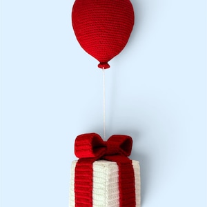 Puede incluir: Decoración hecha a mano con ganchillo que presenta un globo rojo suspendido sobre una caja de regalo blanca con un lazo y una cinta rojos. El globo y la caja de regalo están conectados por una cuerda blanca, creando un diseño festivo y caprichoso.