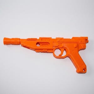 May include: A bright orange plastic toy blaster featuring a long barrel, a textured grip, and a trigger mechanism. The design includes geometric cutouts along the main body and a flared muzzle at the front, presented in a solid, uniform color.
