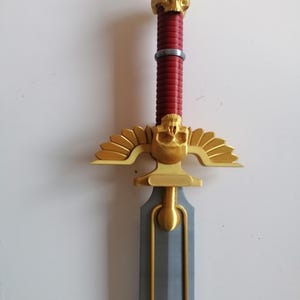 Op de afbeelding: Een decoratief zwaard met een gouden schedel aan de pommel en een gouden pareerstang met gestileerde vleugels. De handgreep is rood en het lemmet is grijs. Het zwaard is een fantasy-thema-item.