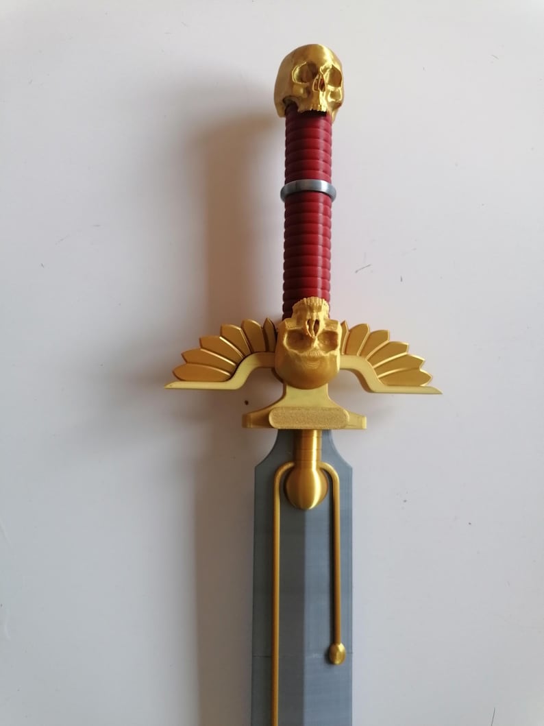 Op de afbeelding: Een decoratief zwaard met een gouden schedel aan de pommel en een gouden pareerstang met gestileerde vleugels. De handgreep is rood en het lemmet is grijs. Het zwaard is een fantasy-thema-item.