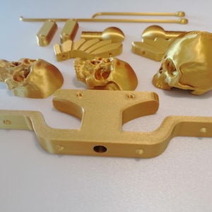 Op de afbeelding: Een verzameling goudkleurige 3D-geprinte onderdelen, waaronder schedelvormige objecten, een gebogen balk en diverse rechthoekige en cilindrische stukken. De componenten zijn gerangschikt op een wit oppervlak, wat wijst op een doe-het-zelf- of knutselproject.