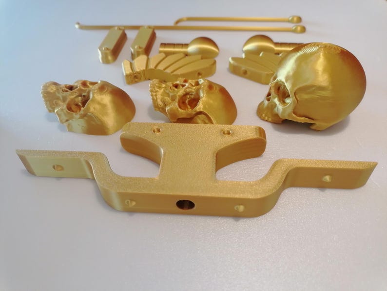 Op de afbeelding: Een verzameling goudkleurige 3D-geprinte onderdelen, waaronder schedelvormige objecten, een gebogen balk en diverse rechthoekige en cilindrische stukken. De componenten zijn gerangschikt op een wit oppervlak, wat wijst op een doe-het-zelf- of knutselproject.