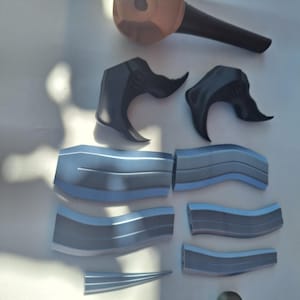 Puede incluir: Un conjunto de piezas impresas en 3D, que incluye una estructura en forma de garra, paneles curvos y una pieza cilíndrica. Las piezas son principalmente grises y azules, con un componente en forma de cabeza de color beige. Los objetos están dispuestos sobre una superficie blanca.