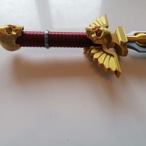 Op de afbeelding: Close-up van een zwaard met een gouden schedel- en vleugelontwerp op de handgreep. De handgreep is bordeauxrood en het lemmet is zilverkleurig. Dit zwaard is waarschijnlijk een verzamelobject of voor cosplay.
