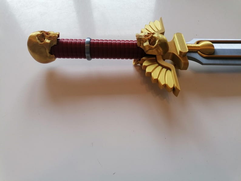 Op de afbeelding: Close-up van een zwaard met een gouden schedel- en vleugelontwerp op de handgreep. De handgreep is bordeauxrood en het lemmet is zilverkleurig. Dit zwaard is waarschijnlijk een verzamelobject of voor cosplay.
