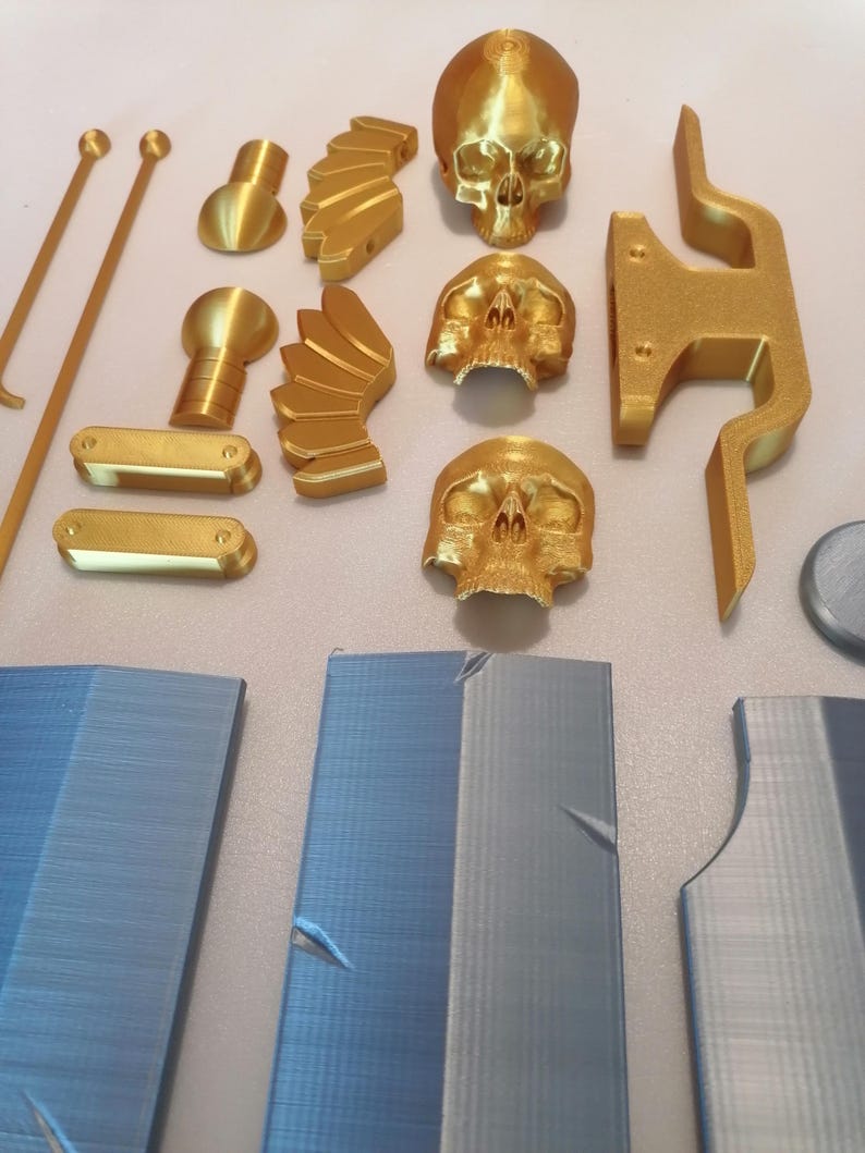 Op de afbeelding: Een verzameling goudkleurige 3D-geprinte objecten, waaronder schedels, decoratieve elementen en componenten voor een grotere structuur. Verschillende blauwgrijze panelen suggereren een project of model in aanbouw.