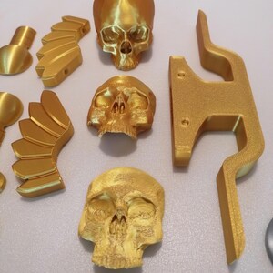 Op de afbeelding: Een verzameling goudkleurige 3D-geprinte objecten, waaronder drie gedetailleerde schedelmodellen, vleugelvormige componenten en een gestileerde beugel. De objecten zijn gerangschikt op een lichtgekleurd oppervlak, waarbij hun ingewikkelde ontwerpen worden getoond.