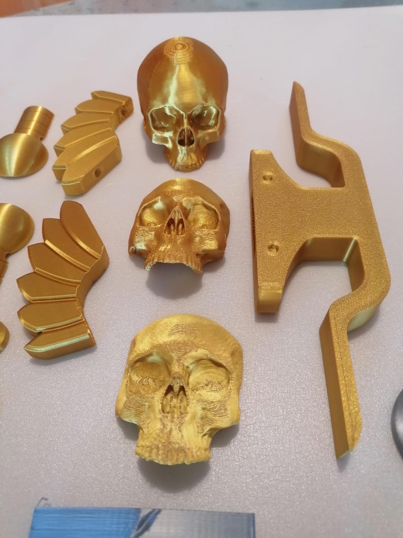 Op de afbeelding: Een verzameling goudkleurige 3D-geprinte objecten, waaronder drie gedetailleerde schedelmodellen, vleugelvormige componenten en een gestileerde beugel. De objecten zijn gerangschikt op een lichtgekleurd oppervlak, waarbij hun ingewikkelde ontwerpen worden getoond.