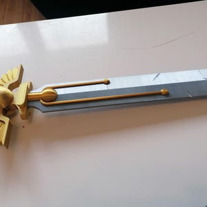 Op de afbeelding: Een gedetailleerd zwaard met een goudkleurig schedel- en vleugelontwerp op de pareerstang. Het lemmet is zilver en grijs, met gouden accenten. De handgreep is omwikkeld met rood materiaal. Een fantasy-item.