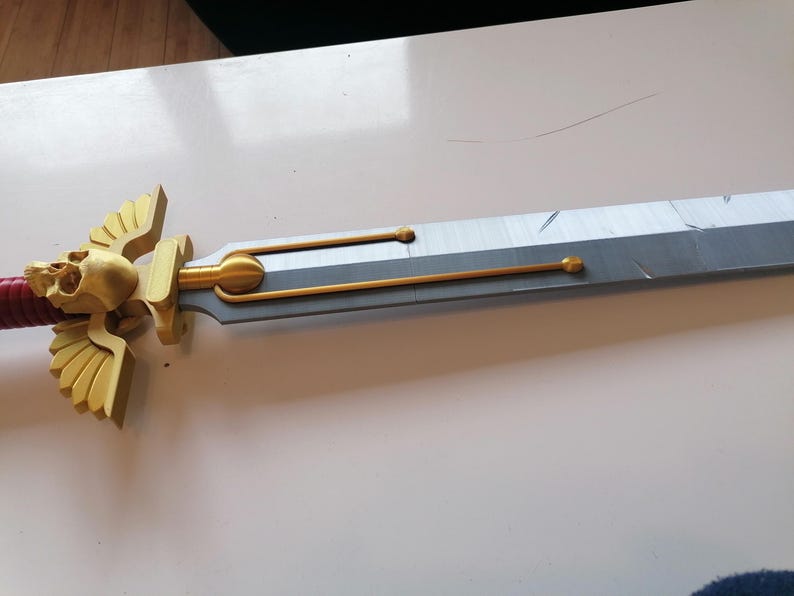 Op de afbeelding: Een gedetailleerd zwaard met een goudkleurig schedel- en vleugelontwerp op de pareerstang. Het lemmet is zilver en grijs, met gouden accenten. De handgreep is omwikkeld met rood materiaal. Een fantasy-item.