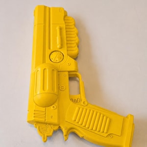 Puede incluir: Un bláster de juguete de color amarillo brillante con un diseño futurista, que presenta un cañón cilíndrico, una empuñadura texturizada y varios detalles mecánicos moldeados, incluyendo rejillas de ventilación circulares y una varilla montada en el lateral. El objeto tiene un acabado mate sólido.