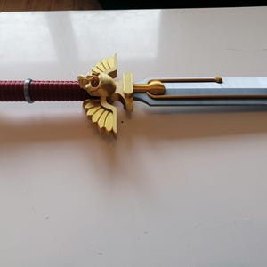 Op de afbeelding: Een gedetailleerd zwaard met een goudkleurige schedel op de pommel en de pareerstang. De handgreep is omwikkeld met rood materiaal. Het lemmet is zilverkleurig met gouden accenten. Fantasy-artikel.